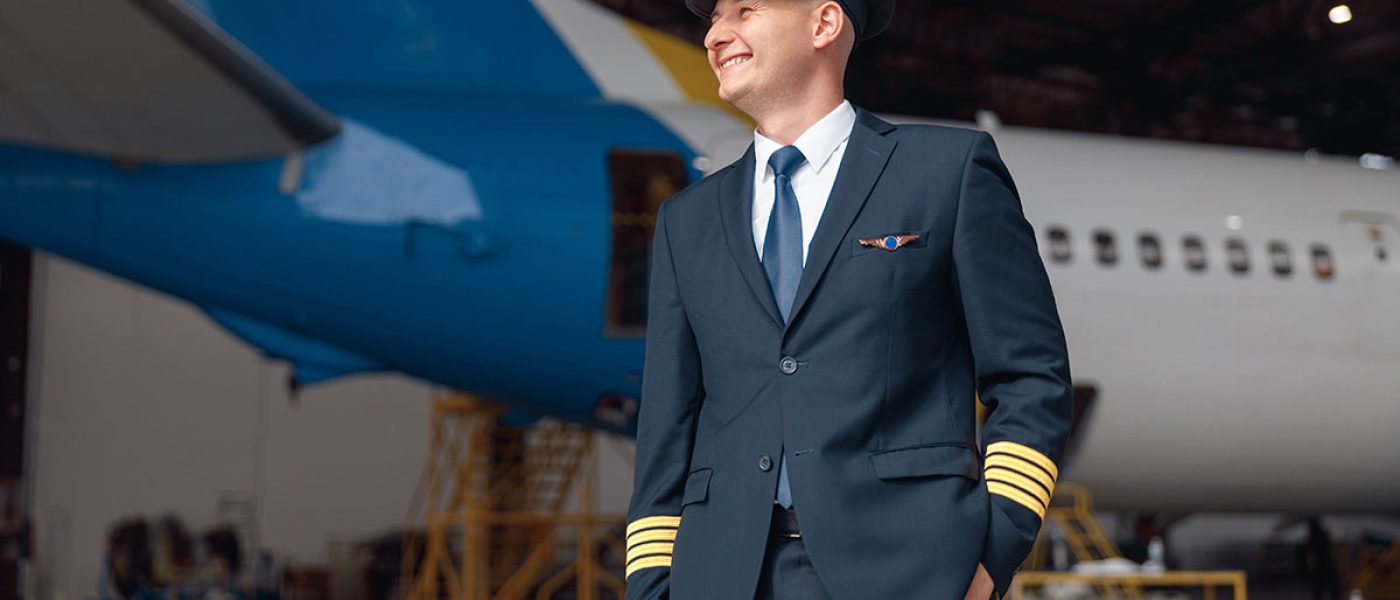 Cheerful pilot in uniform smiling away, standing in front of big passenger airplane in airport hangar. Aircraft, occupation, transportation concept